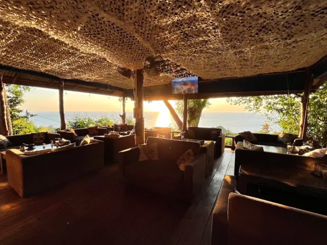 An open wooden lounge with sofas under a woven canopy, sunset over the sea