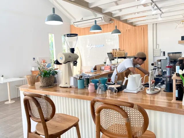 A barista pouring from a kettle at a light wooden counter, a grinder and cane chairs