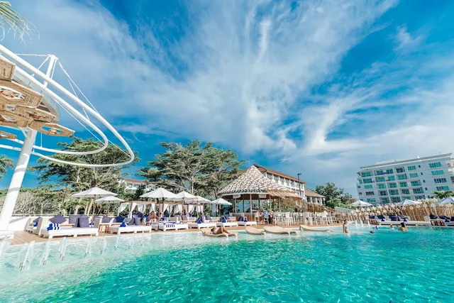 A turquoise pool with fountains, white sunbeds and a thatched structure under blue skies