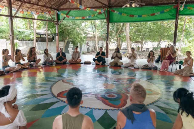A group sitting in a circle on a painted mandala floor, an open pavilion with prayer flags