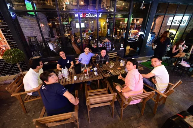 A group sitting around wooden tables on the sidewalk in front of a lit glass frontage in the evening