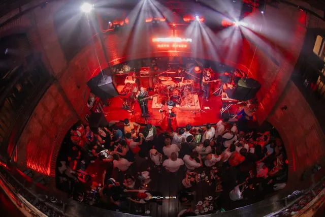 A band playing on a stage beneath a red brick arch, facing a dense crowd below