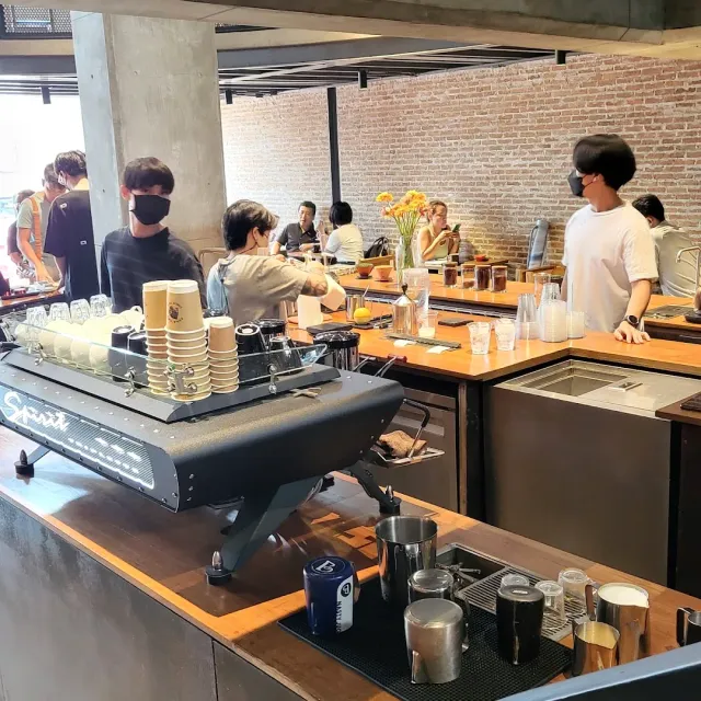 A busy counter with an espresso machine, a stack of cups, baristas and a brick wall