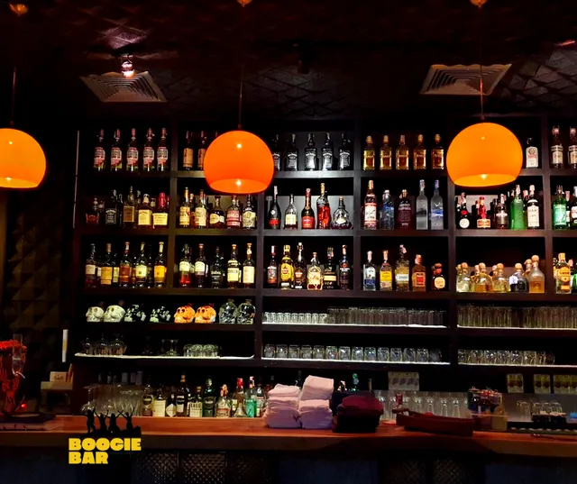 Packed bottle shelves behind a wooden counter, under three hanging orange lamps