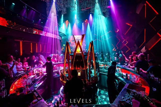 A round bar lit in orange neon in the middle of a club, with purple light beams and a dancing crowd