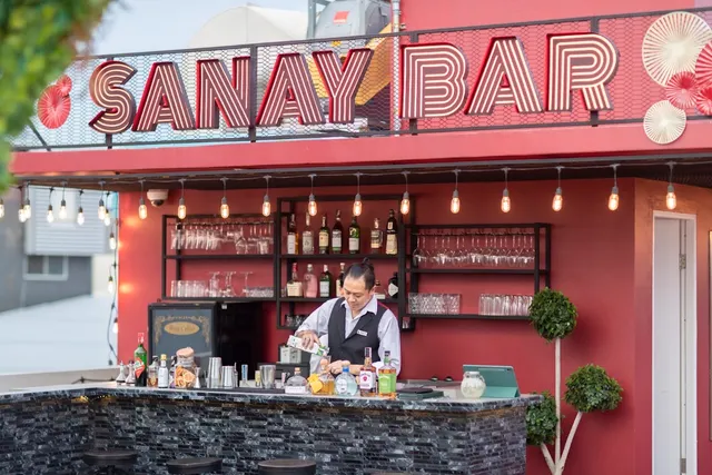 A bartender pouring a drink behind a rooftop counter, under a large letter sign and hanging bulbs