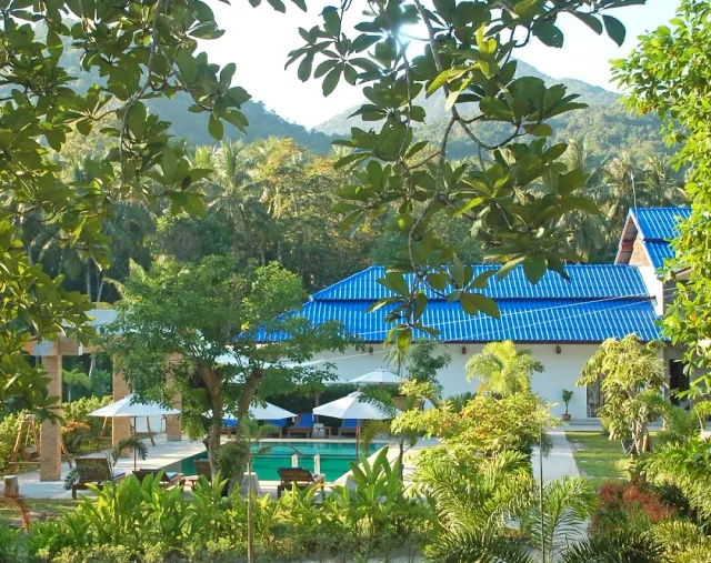 A building with a blue tiled roof and a pool with parasols, forested hills behind