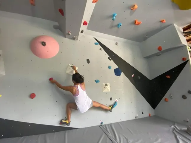 A climber on a gray bouldering wall with colorful holds above a landing mat