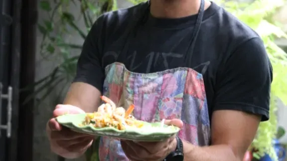 A man in a colorful apron holding a finished dish on a green banana leaf