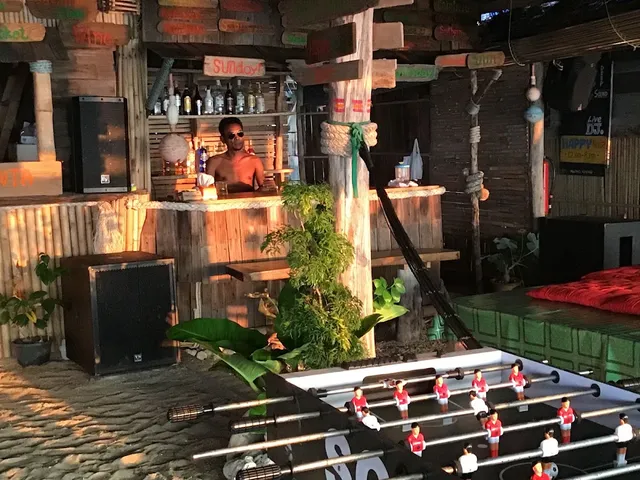 A rustic wooden bar with colorful signs, a bartender and a foosball table on a sand floor