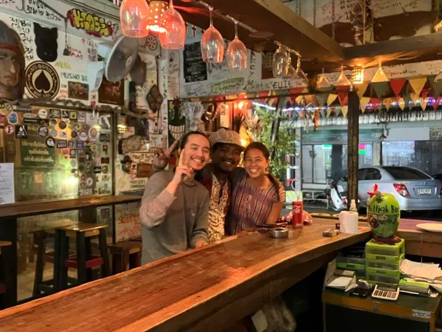 Three people smiling behind a wooden counter, walls covered in stickers and hanging glasses