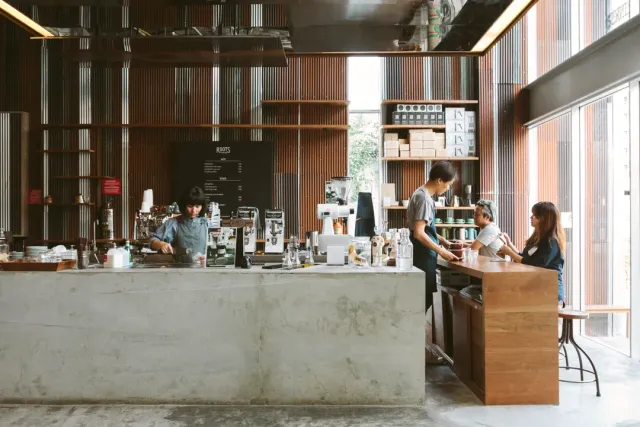 A central concrete counter with two baristas, slatted wooden walls and customers to the side