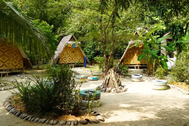 Triangular bamboo huts in a sandy yard, a prepared bonfire and green vegetation around