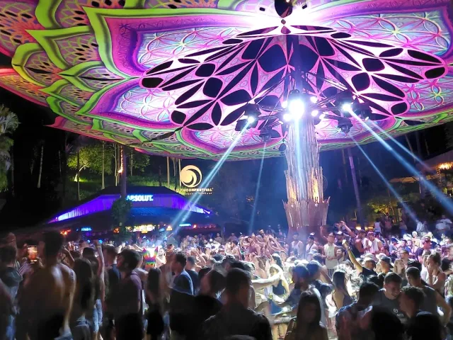 A lit fabric dome in a mandala pattern above a dancing crowd, light beams and treetops