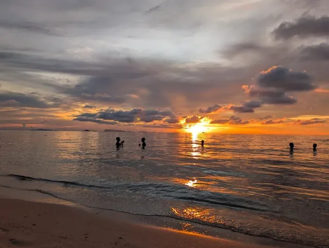 An orange sunset over the sea with silhouettes of bathers standing in the shallow water