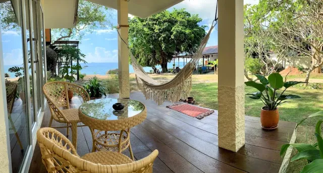 A wooden veranda with a rattan table and chairs, a hanging hammock and a sea view behind