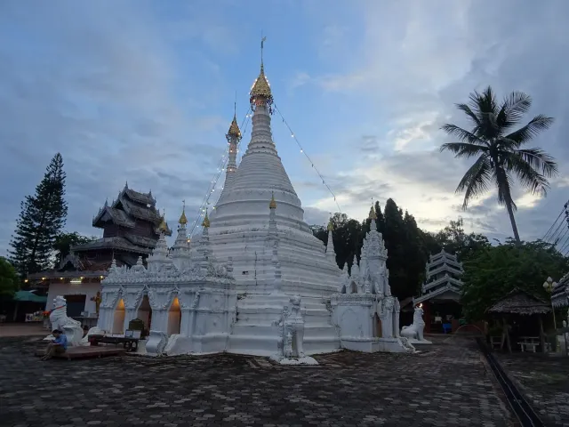 A white pointed chedi on a hilltop beside a wooden hall and palms