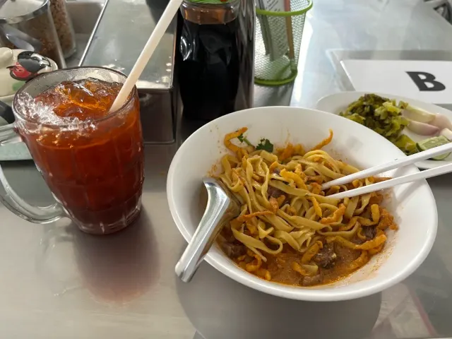 A bowl of curry noodles with a spoon beside a glass of iced tea on a metal table