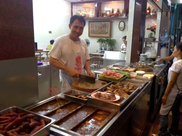 A smiling cook behind an open counter with trays of meat and dark sauce