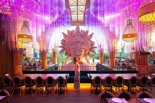 An ornate space with a pink and purple strand curtain, a large face sculpture and bistro chairs