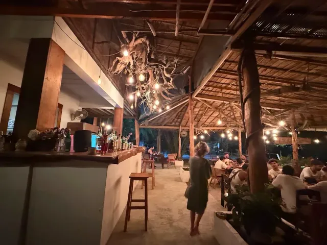 An open space under a thatched roof in the evening, a branch chandelier with bulbs and a bottle counter