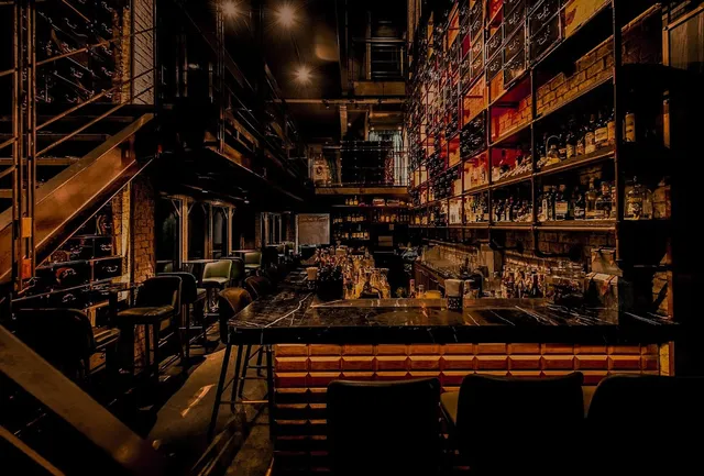 A dim bar with a black marble counter, tall bottle shelves and metal stairs to the side