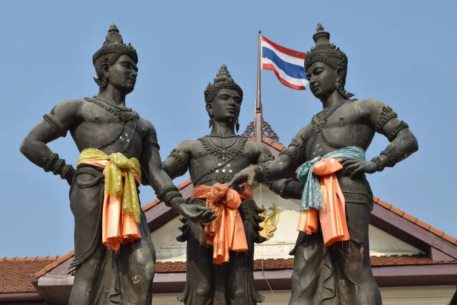 Three bronze statues of standing figures draped in orange cloth beside a flag