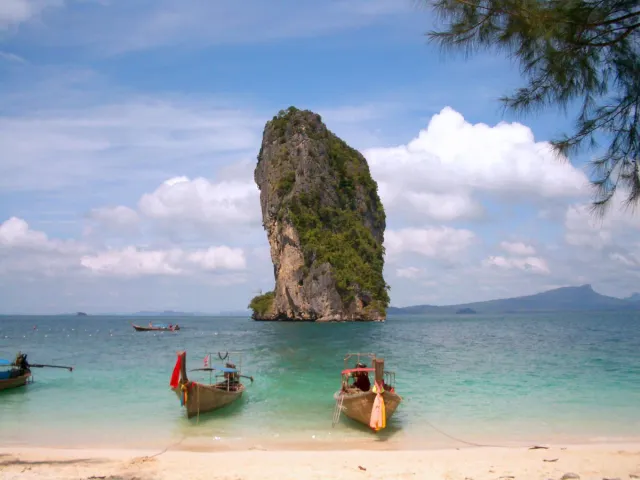 A lone limestone cliff off a white sand beach in turquoise water with long tail boats