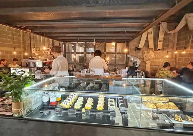 A lit pastry display case in front of a counter, staff behind it and bare block walls