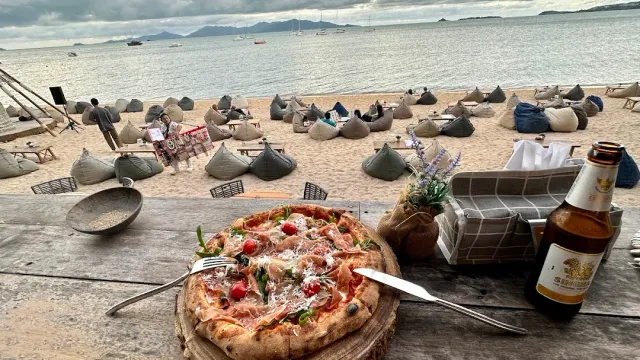 A pizza on a wooden table facing a beach with scattered beanbags and a calm sea