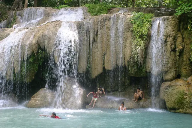 A wide curtain of water falling over a limestone shelf into a pale blue pool with bathers
