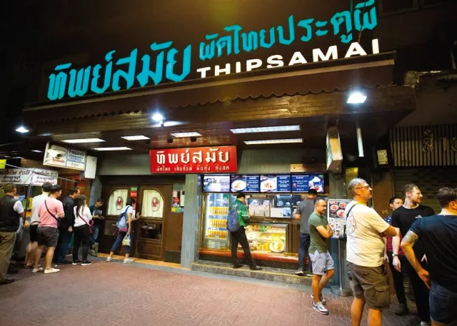 A lit street restaurant front at night with a queue waiting on the sidewalk