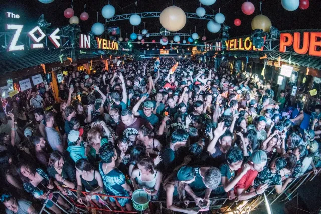A dense crowd in an open venue at night, neon signs and balloons hanging above