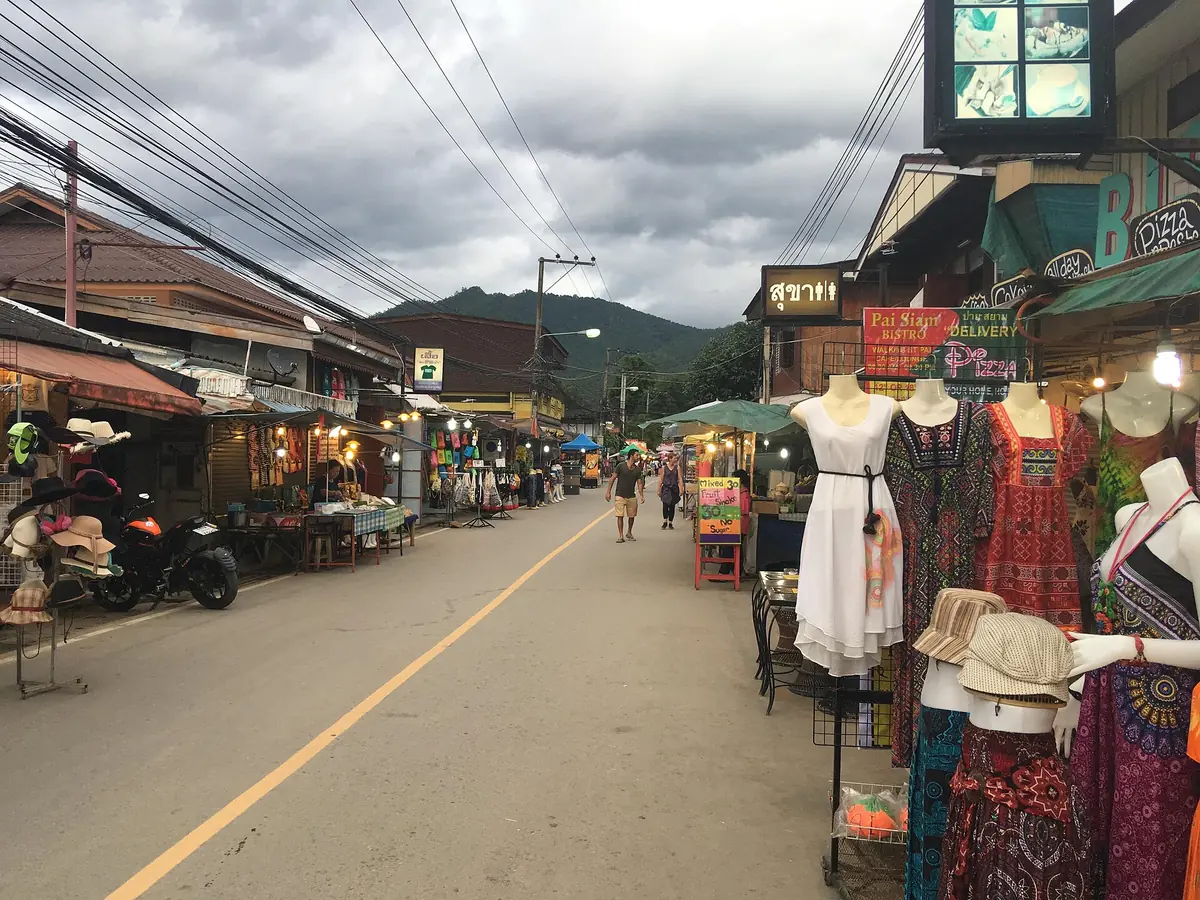A market street with clothing stalls and mannequins on the right, wooden shops on the left and a mountain ridge behind