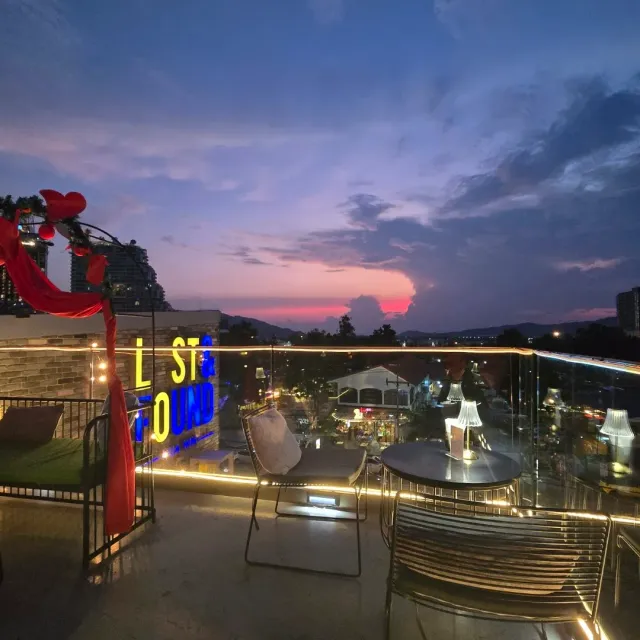 A rooftop terrace at pink sunset with a yellow neon sign, a round table and metal chairs
