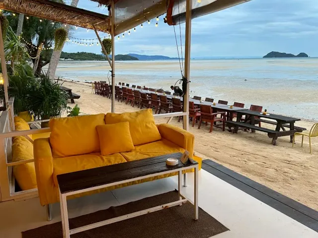 A yellow sofa under a shade and a row of wooden chairs on the sand facing shallow water