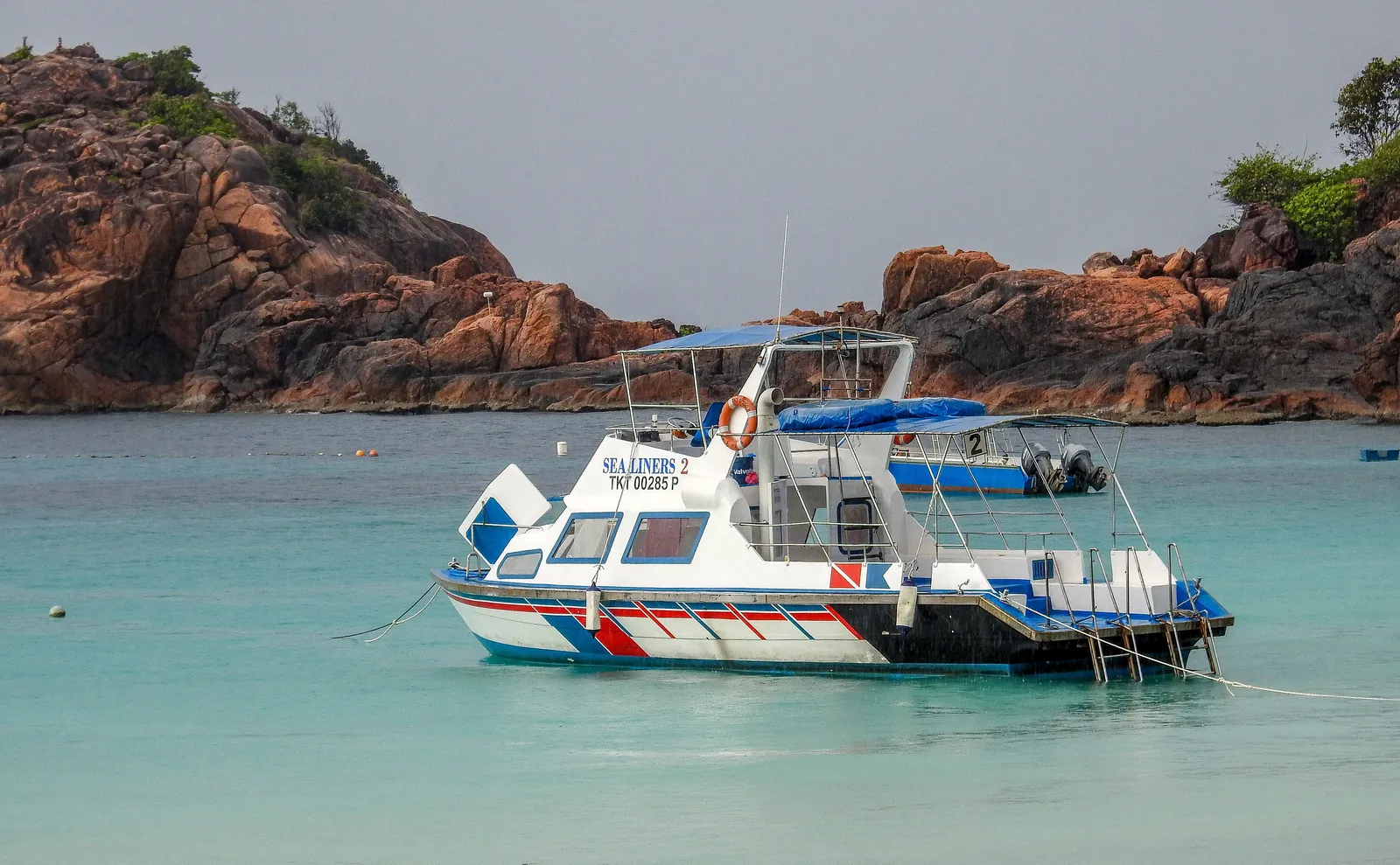 A dive boat anchored beside large granite boulders in clear turquoise water in Thailand