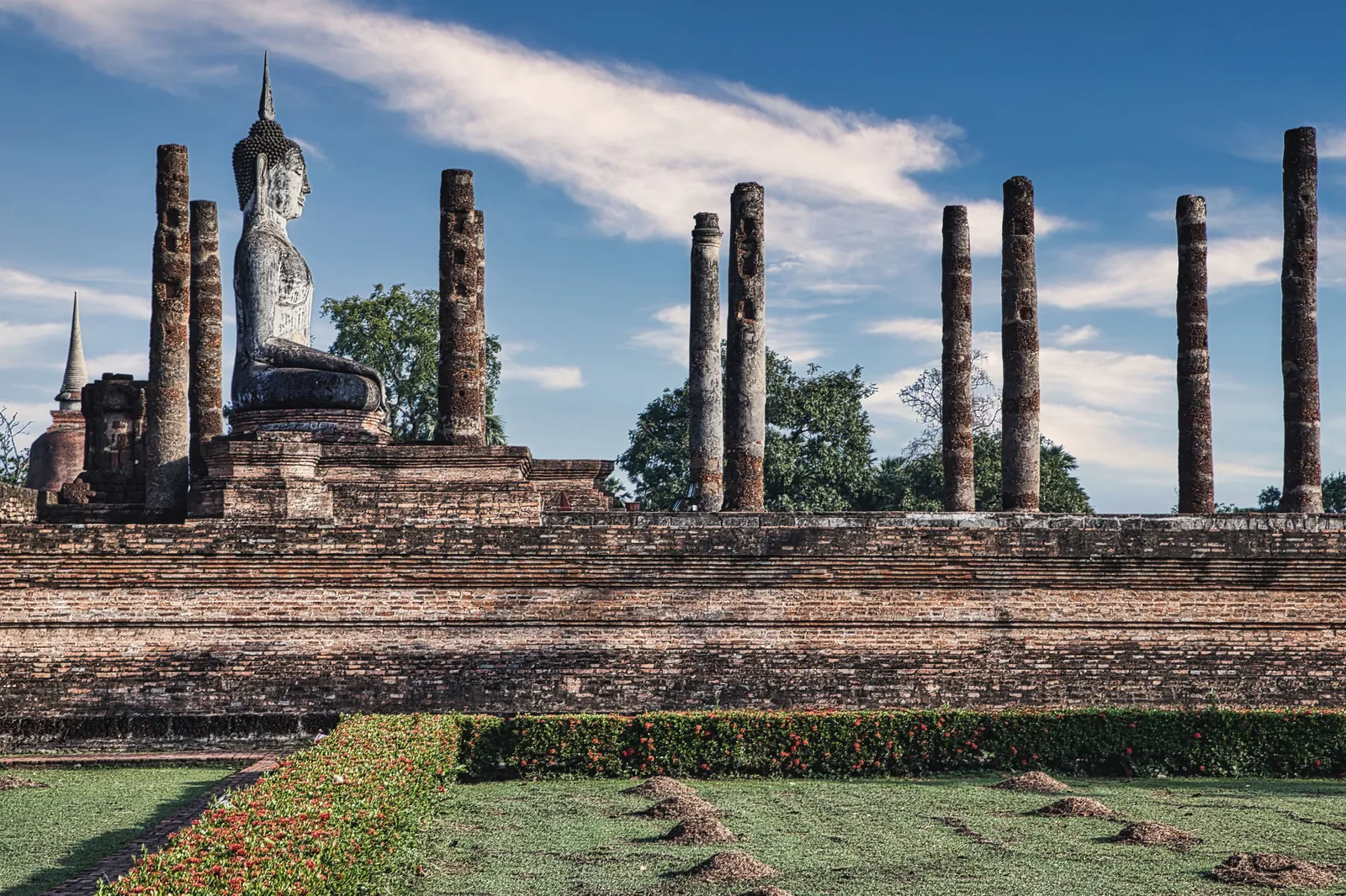 A seated Buddha statue among brick columns and a lotus pond at Sukhothai Historical Park, Thailand