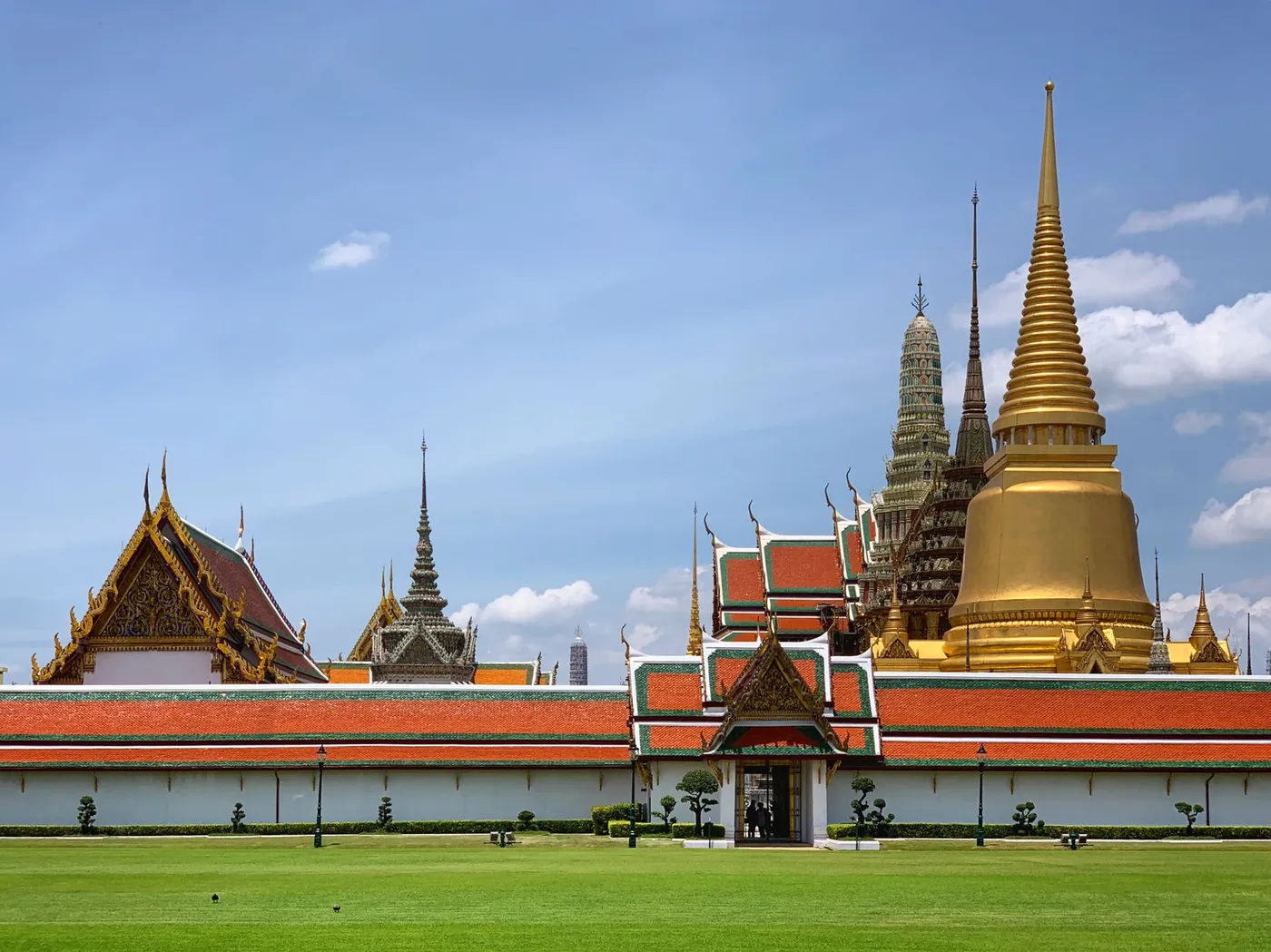 The gold chedi and ornately tiled roofs of Bangkok's Grand Palace complex under a bright blue sky, with a green lawn in the foreground