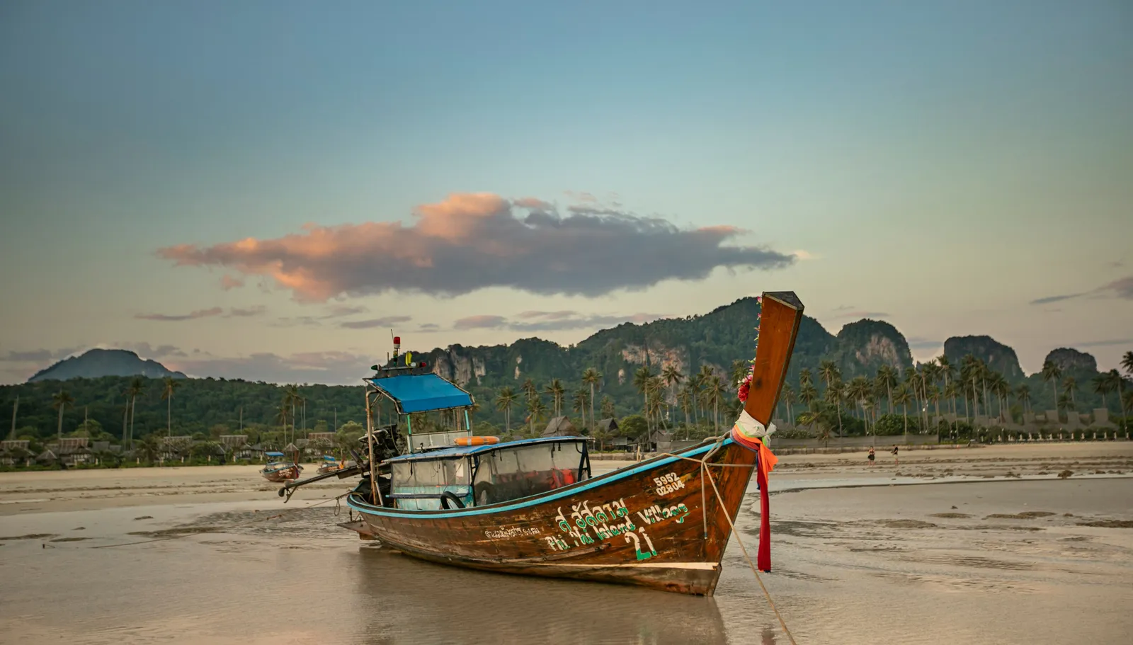 A wooden longtail boat moored off a quiet beach at sunset with limestone hills behind