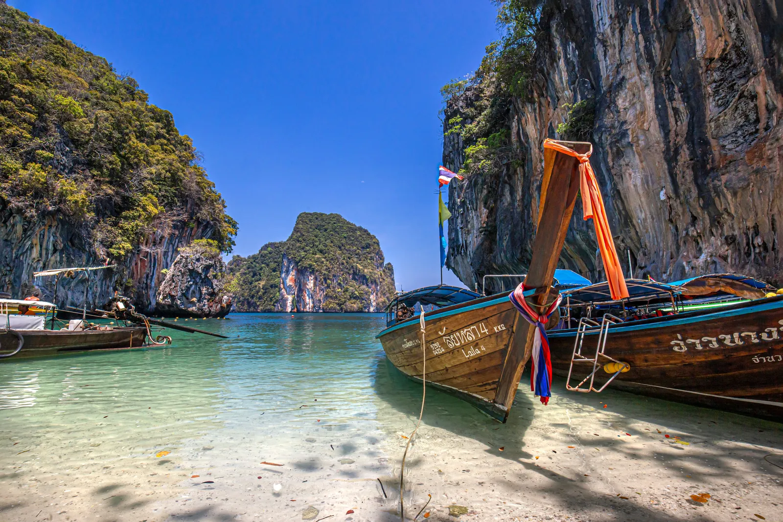 A wooden long-tail boat moored on clear shallow water beside a limestone karst island in the Andaman Sea, Thailand