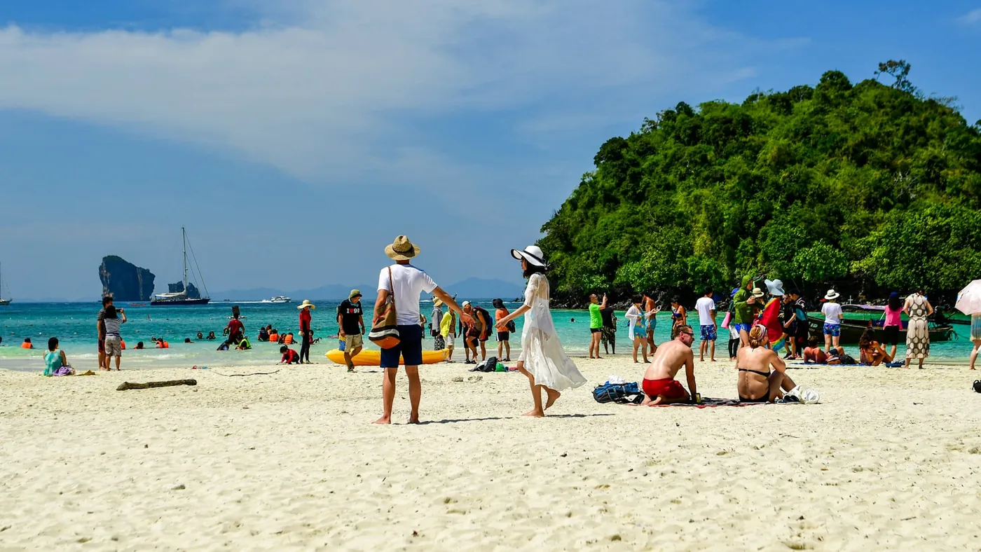 A busy white sand beach crowded with tourists and boats near a green limestone island