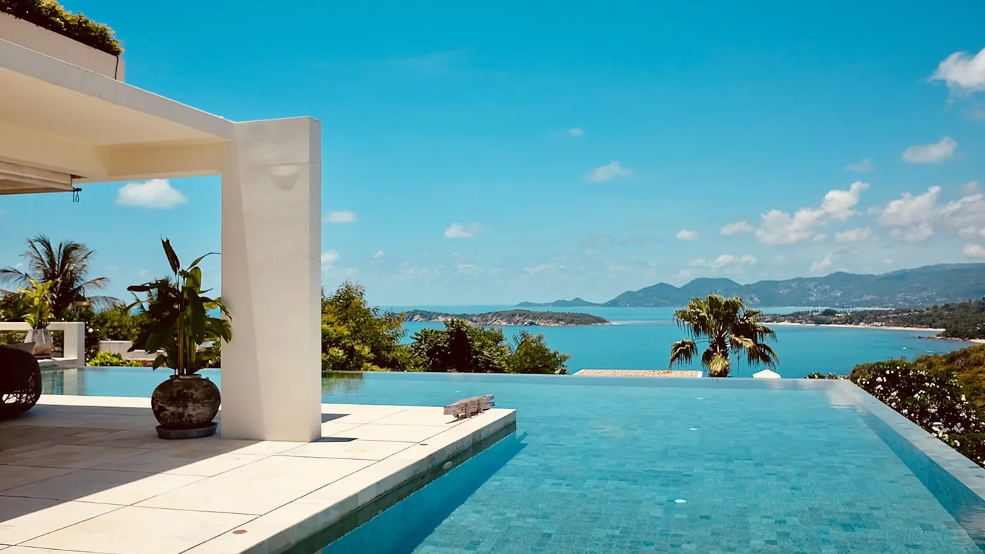 An infinity pool on a resort terrace overlooking a bay and hills on a Thai island, palm trees against a clear daytime sky